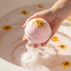 Bombe de bain rose de luxe tenue dans la main, effervescence dans l'eau, enrichie en Beurre de Karité, Absolue de Rose et Encens pour la détente et l'hydratation de la peau.